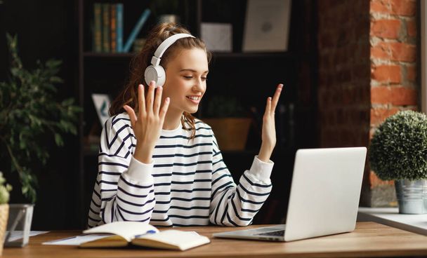 Glückliche Millennial-Frau in lässiger Kleidung und drahtlosen Kopfhörern gestikuliert, während sie Nachrichten mit einem Freund teilt, der Videotelefonie per Laptop am Tisch im Dachgeschoss hat - Foto, Bild