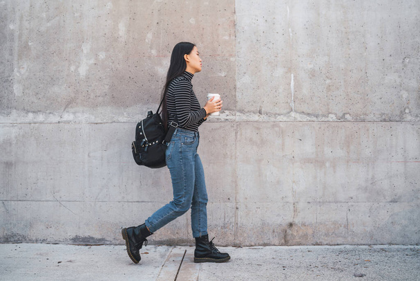 Portrait of young Asian woman walking and holding a cup of coffee against grey wall. - Фото, зображення