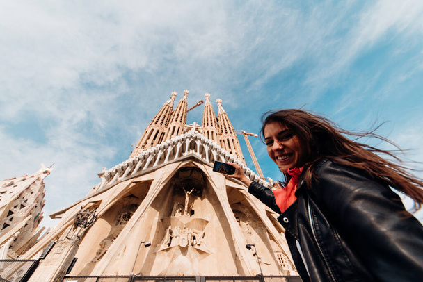 Smiling young woman tourist taking photos with smartphone in front of the famous Sagrada Familia catholic cathedral. Tourism in Barcelona,Spain.Visiting famous landmarks and attractions. - Foto, afbeelding