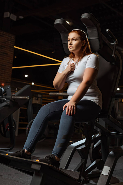 confident overweight girl looking away while sitting on fitness machine - Фото, зображення