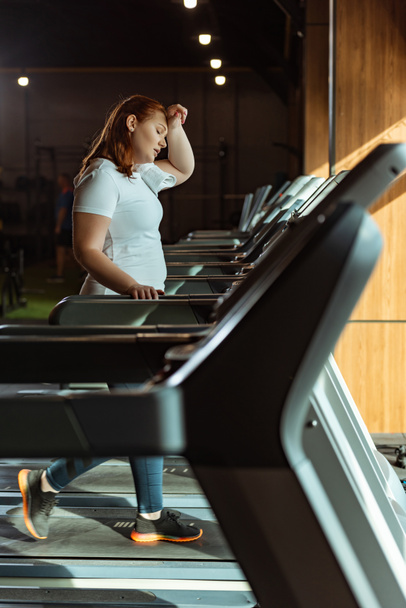 selective focus of tired overweight girl touching forehead while running on treadmill - Fotó, kép