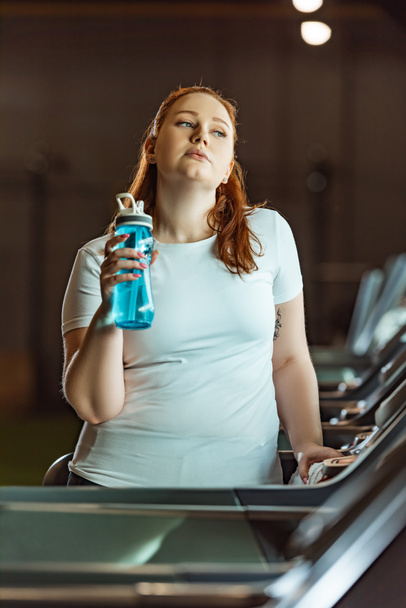 κουρασμένο υπέρβαρο κορίτσι κρατώντας αθλητικό μπουκάλι, ενώ στέκεται στο διάδρομο στο γυμναστήριο - Φωτογραφία, εικόνα