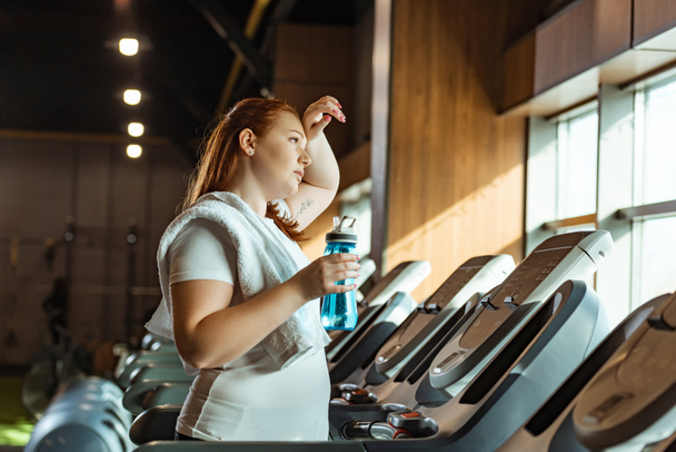 cansada chica con sobrepeso tocando la frente mientras sostiene la botella de deportes mientras está de pie en la cinta de correr
 - Foto, imagen