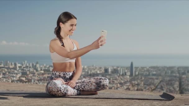 Sportsoman robi selfie na smartfonie w mieście. Joga kobieta siedzi w pozycji lotosu - Materiał filmowy, wideo