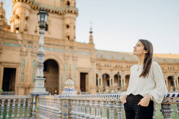 Enjoying beautiful historical landmark.Young tourist woman looking over Plaza de Espana in Seville,Andalusia,Spain.Traveling to Spain.Sunset on Spain Square.Pensive woman in public square in Europe. - 写真・画像