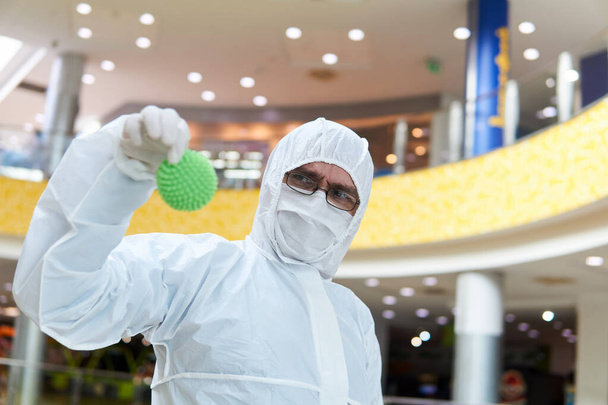 man in coverall disposable anti-epidemic antibacterial isolation suit suspiciously holds in his hand a rubber ball resembling a macro photo of a virus, against the background of some interior of public space - Photo, Image