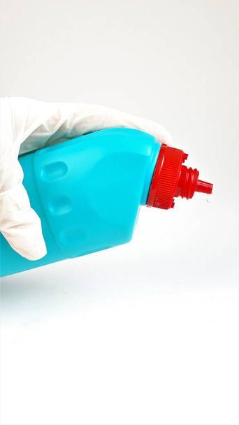 Niebieska plastikowa butelka z czerwonym wieczkiem w dłoni w gumowej białej rękawiczce na białym tle.Płyn do czyszczenia i dezynfekcji pomieszczeń i domów.Zbliżenie. - Zdjęcie, obraz
