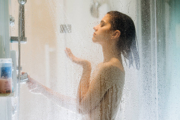 Morning shower.Taking rejuvenating cold shower.Self care moment.Everyday personal hygiene.Unfocused woman showering in glass shower with strong pressure water stream.Focus on drops - 写真・画像