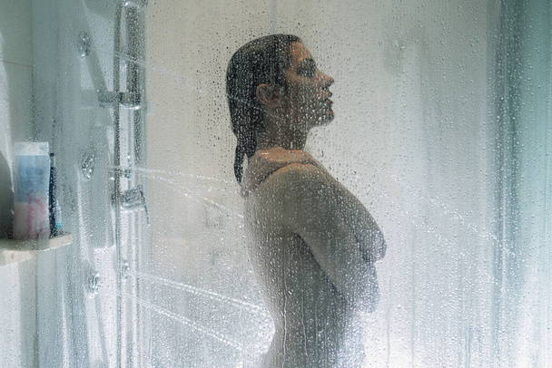 Night shower.Taking relaxing massage shower.Self care moment.Personal hygiene,spa.Unfocused woman showering in glass shower with strong pressure water stream.Focus on drops - Photo, Image