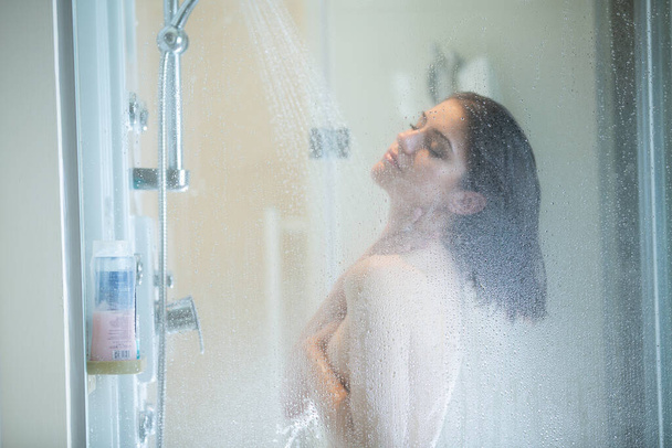 Morning shower.Taking rejuvenating cold shower.Self care moment.Everyday personal hygiene.Unfocused woman showering in glass shower with strong pressure water stream.Focus on drops - Fotografie, Obrázek