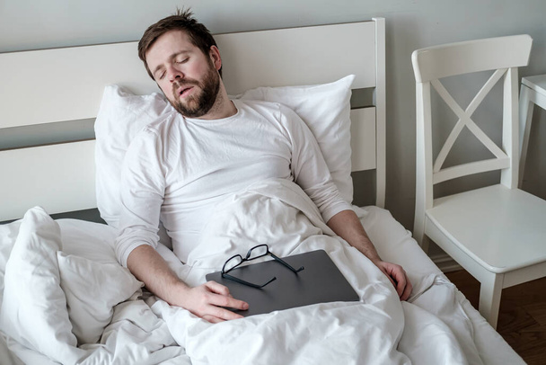 Man was very tired and fell asleep in bed with an ultrabook after he finished working remotely. Concept of quarantine. - 写真・画像