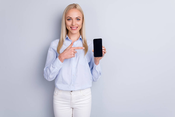 Portrait of positive cheerful promoter girl hold new smartphone device point index finger suggest select wear good look trousers isolated over gray color background - Φωτογραφία, εικόνα