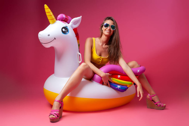 Zomer Mode mooie vrouw in de zomer kleding hebben plezier, glimlachen en poseren met ballonnen Op Unicorn Float op geïsoleerde roze achtergrond. Zomer modieus meisje, roze en vrolijk humeur. - Foto, afbeelding