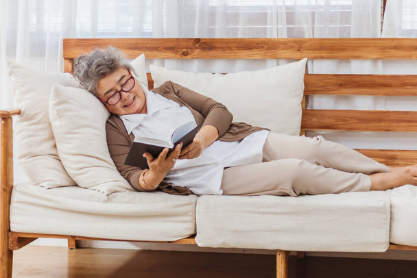 Щаслива і здорова жінка, яка посміхається старшій жінці або пенсіонерці, що читає книжку, відпочиває або сидить на дивані або дивані вдома. Старенький дорослий активний спосіб життя і діяльність у відпустці. Страхування і здоров "я - Фото, зображення