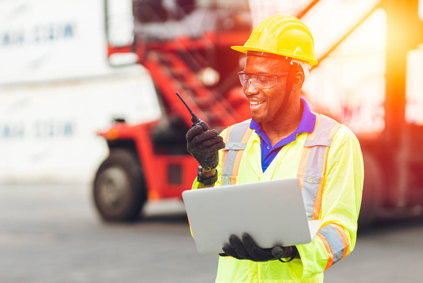 Czarny afrykański szczęśliwy pracownik pracujący w komunikacji logistycznej za pomocą radia i laptopa do kontroli kontenerów załadunkowych w porcie ładunków do samochodów ciężarowych na eksport i import towarów. - Zdjęcie, obraz
