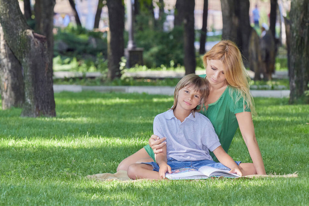 Reading on the lawn. Beautiful mother and cute son read a book on a green grassy meado - Photo, Image
