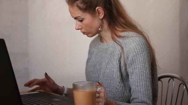 Κορίτσι πληκτρολογώντας σε ένα φορητό υπολογιστή, ενώ κάθεται σε ένα τραπέζι με ένα φλιτζάνι καφέ. Καραντίνα. Βίντεο - Πλάνα, βίντεο