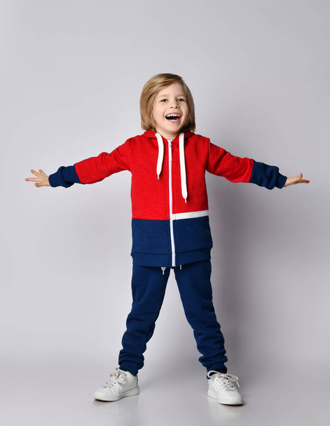 Happy screaming singing blond kid boy in blue and red sportswear hoodie and pants stands with his hands wide apart - Fotografie, Obrázek