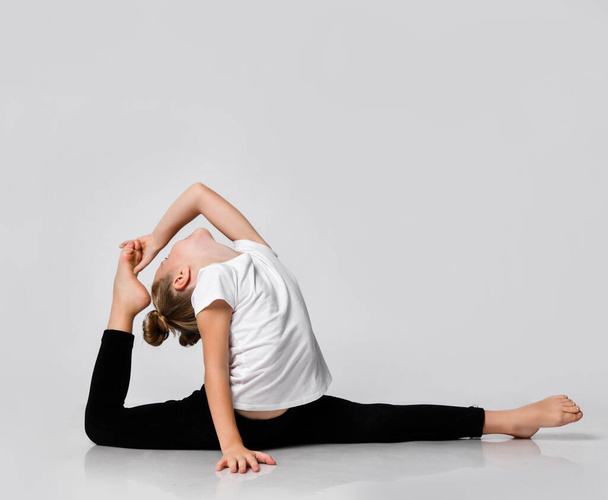 Kid girl is doing gymnastic exercises at home in studio splits with one leg up body bent and head thrown back to foot - Photo, image