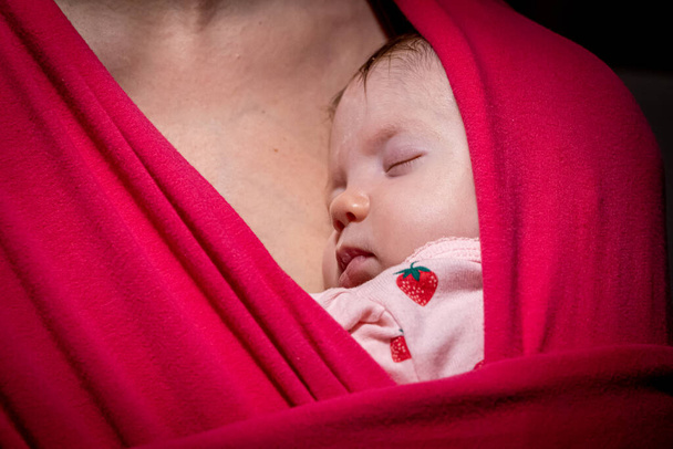 Anya parittyában hordja az aranyos kislányát. Az újszülött egy parittyában alszik, az anyja ölelésében. A kislány 2 hónapos.. - Fotó, kép