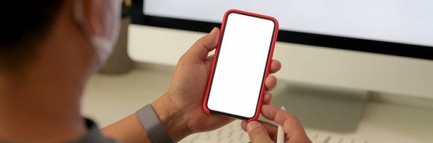 Captura recortada de un hombre usando un teléfono inteligente de pantalla en blanco mientras está sentado en el lugar de trabajo con un dispositivo informático
 - Foto, Imagen
