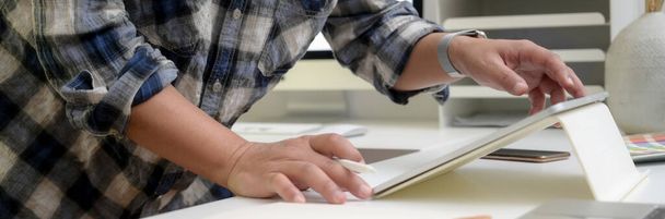 Imagen recortada del diseñador masculino de pie y trabajando en la tableta digital en la mesa blanca en la sala de oficina
 - Foto, imagen
