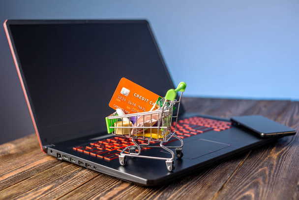 クレジットカードのあるショッピングカートは、青い背景のスクリーンの前にラップトップに立っています。現代の生活の中でインターネット上でオンライン購入の概念 - 写真・画像
