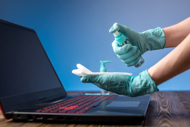 Τα χέρια των γυναικών σε αποστειρωμένα γάντια χειρίζονται το χώρο εργασίας τους σκουπίζοντας μια πετσέτα με ένα απολυμαντικό φορητό υπολογιστή. Καθαριότητα και υγιεινή στην καταπολέμηση των ιών - Φωτογραφία, εικόνα