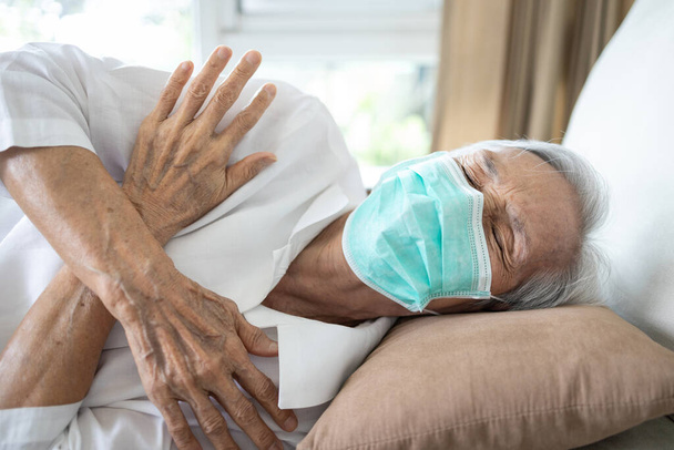 Άρρωστη ηλικιωμένη γυναίκα που φοράει μάσκα έχει πυρετό, ρίγη, υψηλή θερμοκρασία, ψύχρα, οι ηλικιωμένοι έχουν κρυολόγημα, γρίπη, κουρασμένοι ηλικιωμένοι ασθενείς με μυϊκό πόνο σε όλο το σώμα, πόνους από πυρετό, λοίμωξη από τον ιό του κερατοειδούς - Φωτογραφία, εικόνα