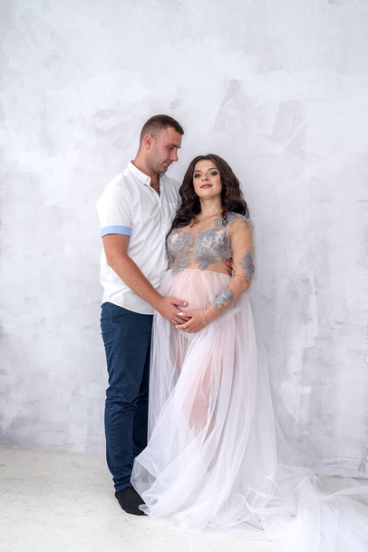 Glückliches und attraktives schwangeres Paar posiert im Studio. Ehemann zärtlich umarmt seine Frau - Foto, Bild