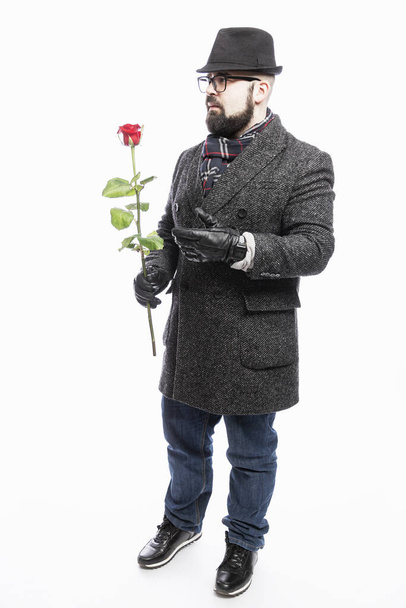 Stylový muž v kabátě a klobouku s rudou růží v ruce. První rande. Bílé pozadí. Svisle. - Fotografie, Obrázek