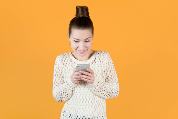 girl enthusiastically looks at the phone, chatting with someone perhaps, Isolated on orange-yellow - Фото, изображение