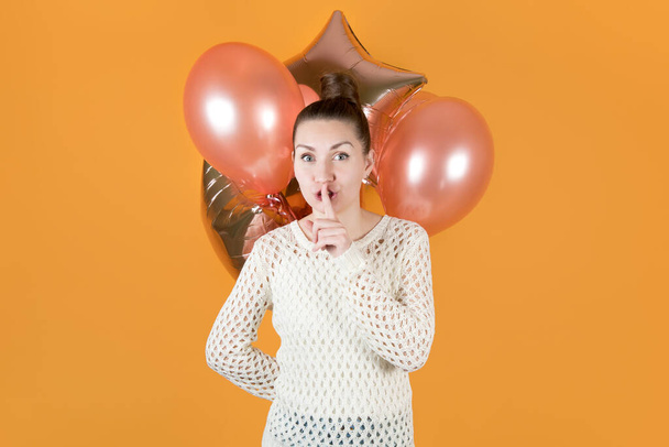 girl covers her mouth with a finger urging to be silent and holds festive balloons behind her. isolated on orange-yellow - Valokuva, kuva