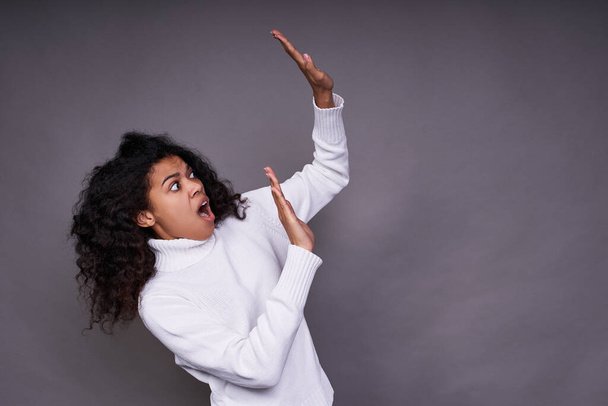 Frightened, troubled young Afro woman, in a white sweater, with curly curly hair, raised her hands up, something falls on her, she defends herself. Posing on a gray background. - Photo, Image