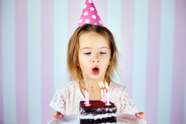 Dziewczynka w różowej czapce zdmuchiwała świeczki na urodzinowym torcie czekoladowym w domu. Portret dziecka urodzinowego. - Zdjęcie, obraz