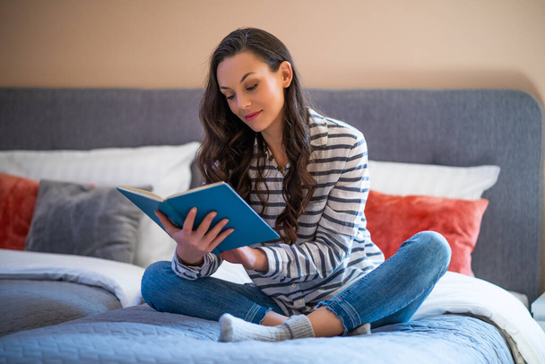 Ευτυχισμένη νεαρή γυναίκα που κάθεται στο κρεβάτι στο σπίτι της και διαβάζει βιβλίο. - Φωτογραφία, εικόνα