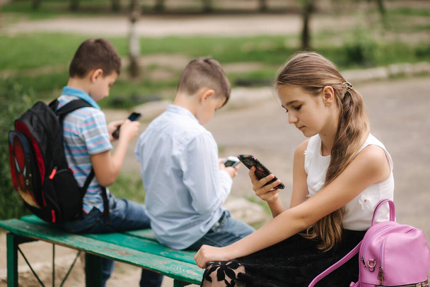 Dva kluci a holka používají telefony během školních přestávek. Roztomilí kluci sedí na lavičce a hrát online hry - Fotografie, Obrázek