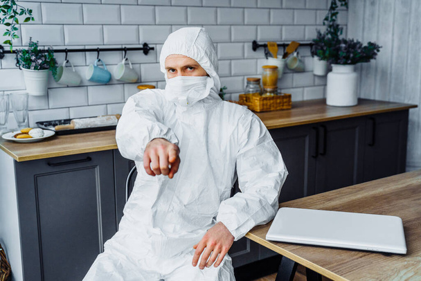 Ο άνθρωπος με το ιικό προστατευτικό λευκό κοστούμι και τη μάσκα προσώπου κάθεται στο σπίτι με τον υπολογιστή και δείχνει προς την κάμερα με ένα δάχτυλο. Απομονωμένο λόγω επιδημίας του κορωναϊού και καραντίνας. - Φωτογραφία, εικόνα