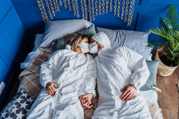 Δύο άνθρωποι με προστατευτικές στολές και μάσκες προσώπου, ξαπλωμένοι στο κρεβάτι και φιλιούνται μέσα από μάσκες. Απομονωμένος στο σπίτι λόγω καραντίνας για προστασία λόγω του ιού της στέψης. Η ασφάλεια προηγείται. - Φωτογραφία, εικόνα