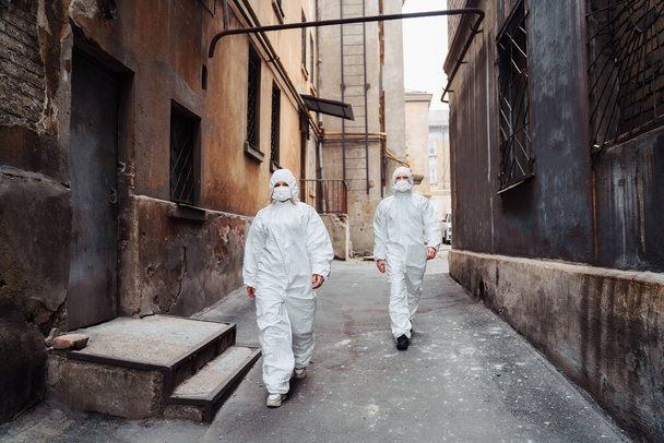 保護服やマスクを着た人々は、戦争中に捨てられた、放棄された、荒廃した通りに沿って屋外を歩いています。コロナウイルスの概念. - 写真・画像