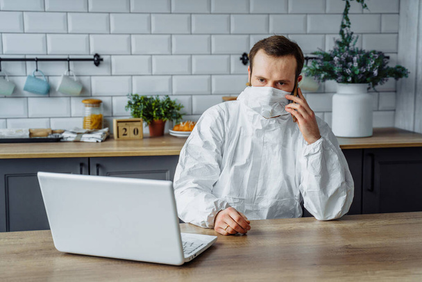 Portrait d'un homme européen portant une combinaison de protection blanche et un masque facial, travaillant de la maison avec un ordinateur portable et un téléphone. Quarantaine due à l'épidémie de coronavirus et isolée de la ville
 - Photo, image
