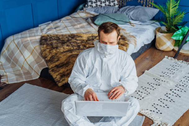 白いウイルス保護衣装を着た白人男性とベッドの近くに座って、コロナウイルスのためにラップトップで自宅から働いている顔のマスクの肖像画。隔離のために自宅で隔離｜covid-19 - 写真・画像