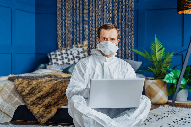 Retrato de homem caucasiano em traje protetor viral branco e máscara facial sentado perto da cama e trabalhando de casa no laptop por causa do coronavírus. Isolado em casa devido à quarentena covid-19
 - Foto, Imagem