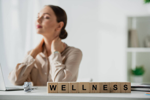 selective focus of businesswoman resting at workplace with laptop and alphabet cubes with wellness word  - Фото, изображение