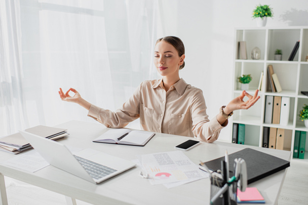 femme d'affaires heureuse méditant avec les yeux fermés et gyan mudra sur le lieu de travail avec ordinateur portable et smartphone
 - Photo, image