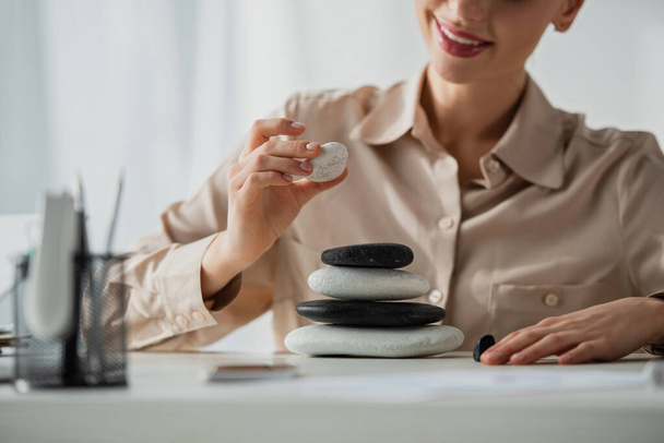 abgeschnittene Ansicht einer glücklichen Geschäftsfrau, die am Arbeitsplatz mit Zen-Steinen sitzt  - Foto, Bild