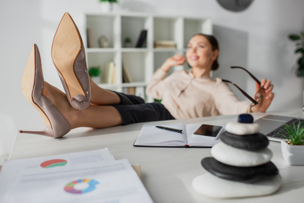 selectieve focus van zakenvrouw zitten met voeten op tafel met zen stenen in het kantoor - Foto, afbeelding