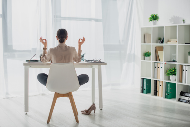 Vista posterior de la mujer de negocios meditando en pose de loto con gyan mudra en el lugar de trabajo
  - Foto, Imagen