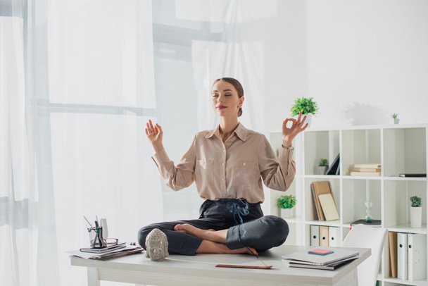 atractiva empresaria meditando en pose de loto con gyan mudra en el lugar de trabajo con cabeza de buda y varilla de incienso
 - Foto, Imagen