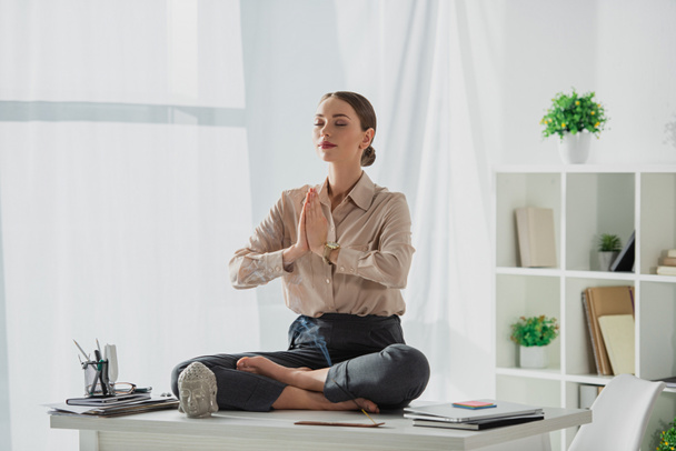 empresaria corporativa meditando en pose de loto con gesto namaste en el lugar de trabajo con cabeza de buda y varilla de incienso
 - Foto, Imagen